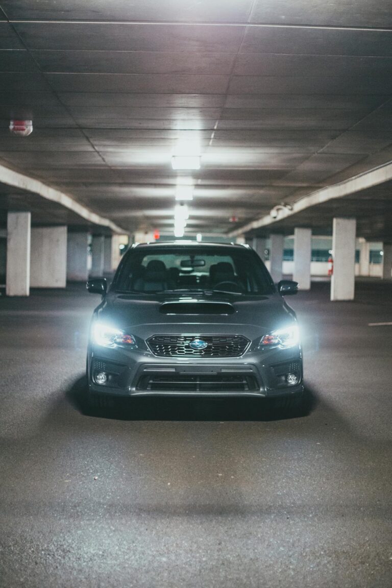 Gray expensive automobile with glowing headlights parked on asphalt in underground parking lot under ceiling with lamps
